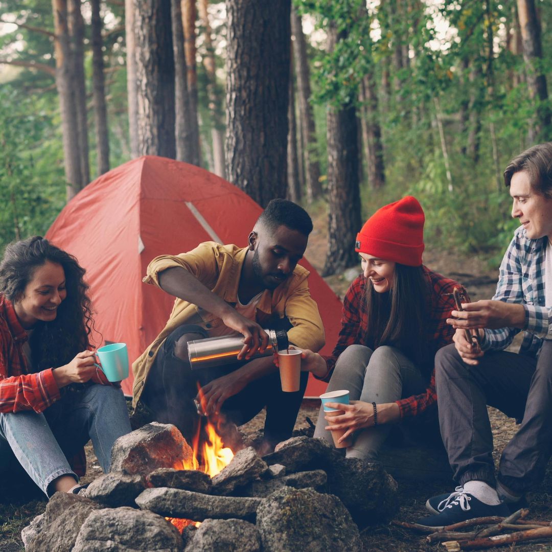 Group of people camping and sitting around a fire with a red tent in the background.