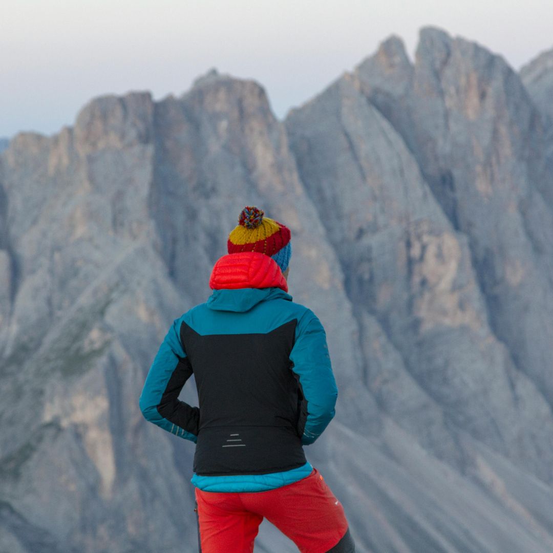 Person wearing a blue and black jacket with red pants and a colorful beanie, standing in front of mountainous landscape.