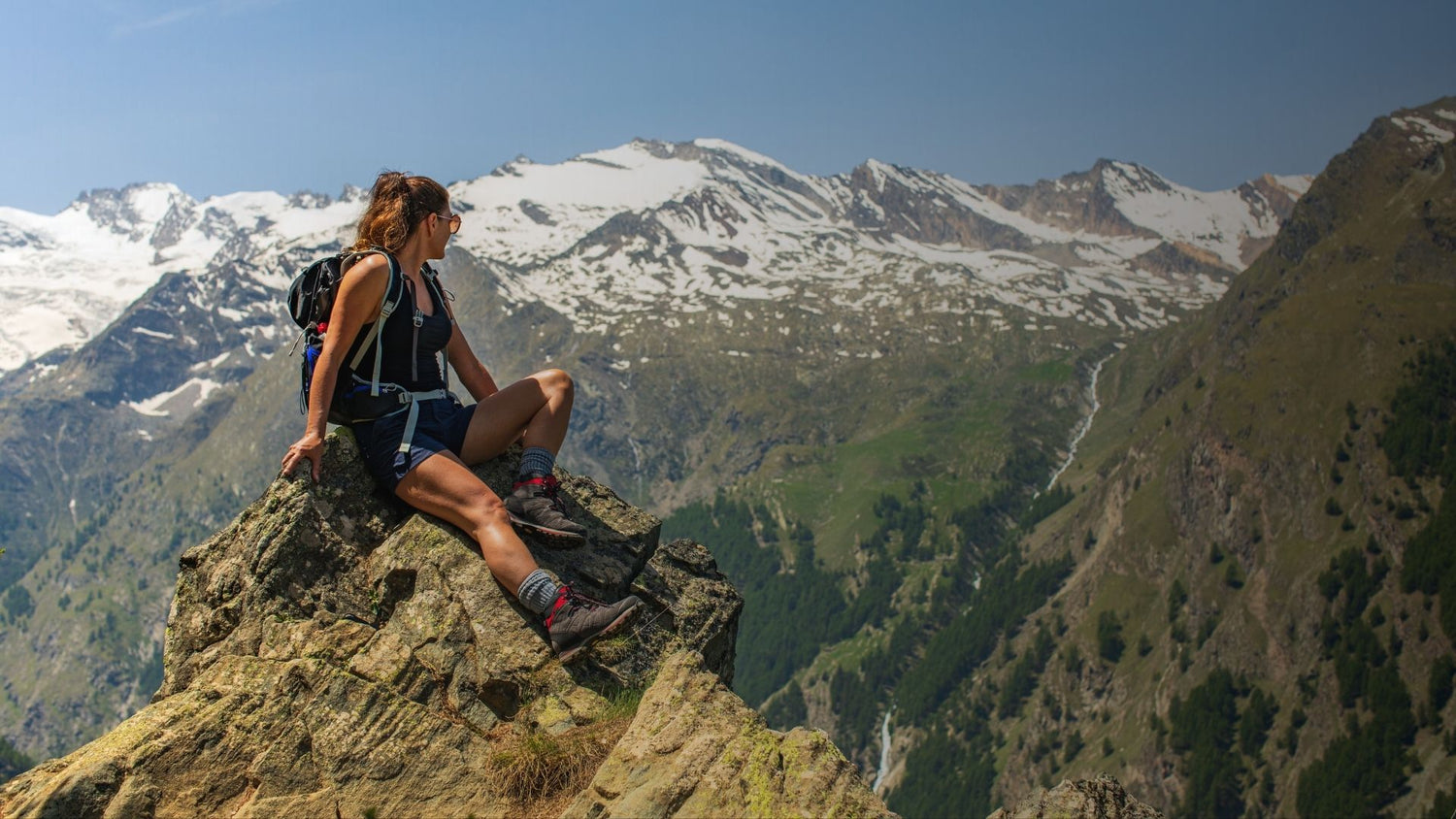 Person sitting on a mountain peak with snow-capped mountains in the background