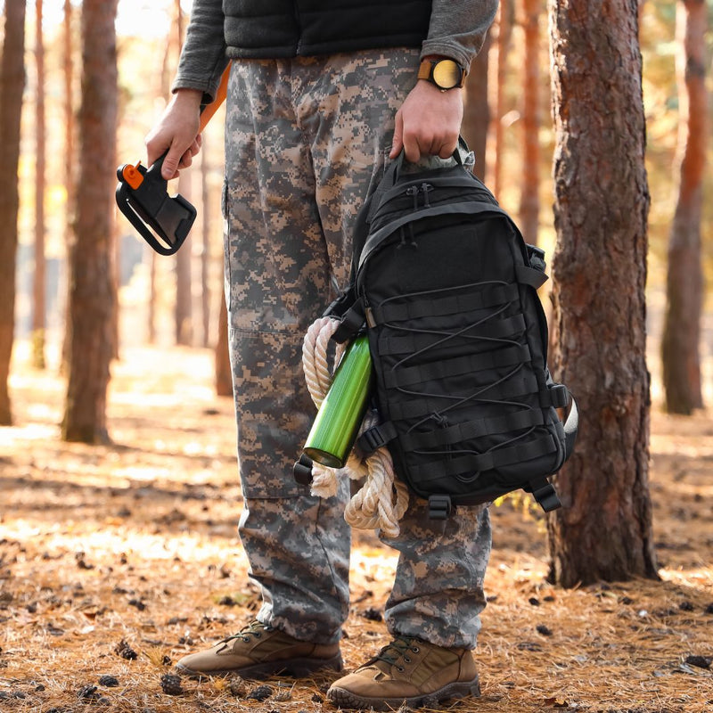Person in camouflage pants and boots holding a black backpack with a green object in a forest setting.